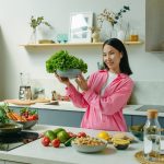 Woman in a kitchen holding fresh lettuce, surrounded by healthy ingredients like tomatoes and avocados, promoting healthy eating.